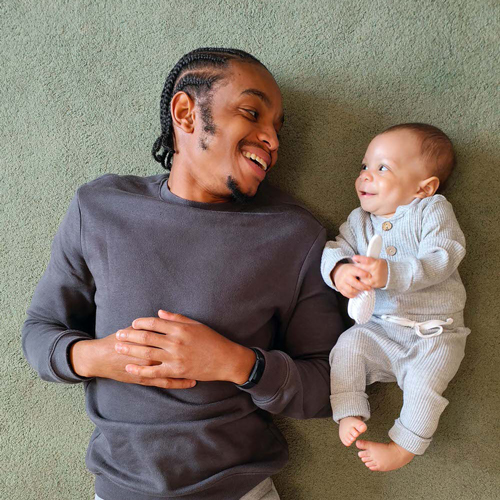 African American man and baby laying on the floor on their backs smiling at each other